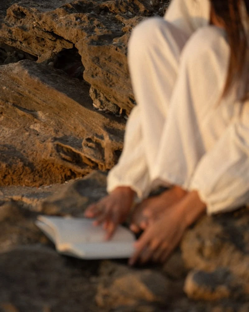 Woman in white linen sitting on sun-warmed rocks with an open book, softly out of focus