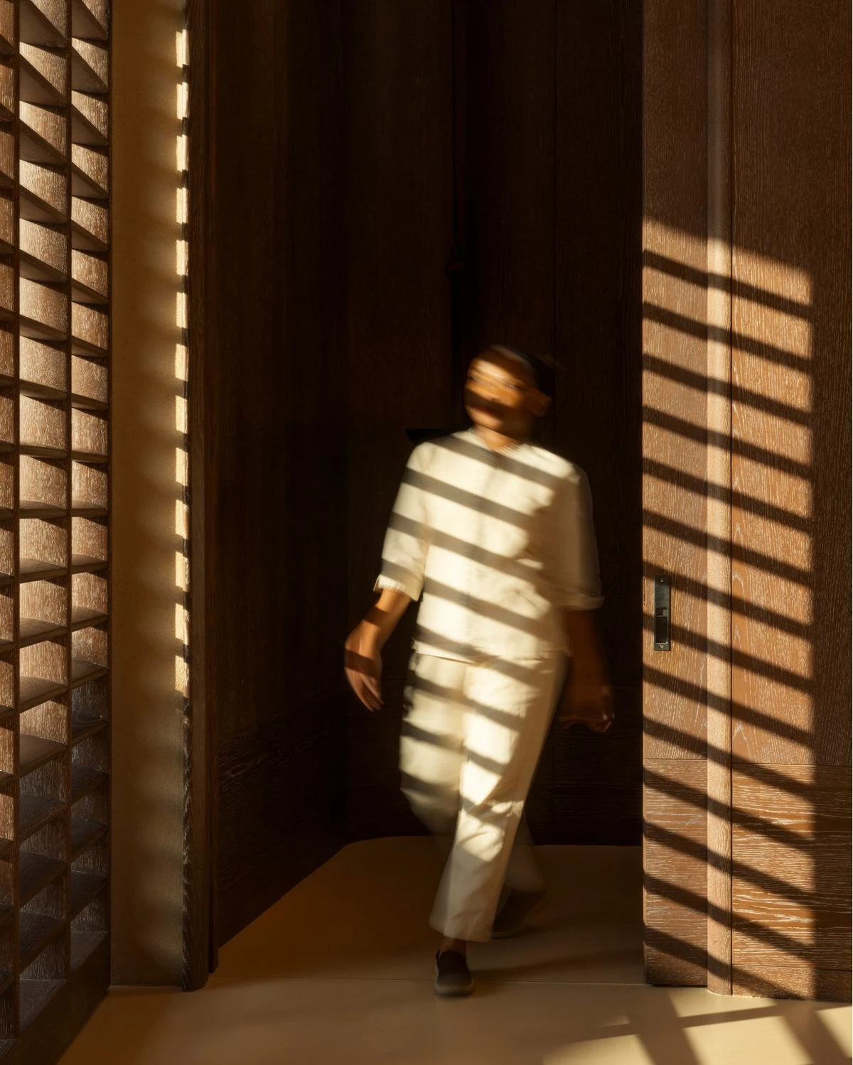 Person in white linen walking through a wooden corridor, striped with warm sunlight through shutters